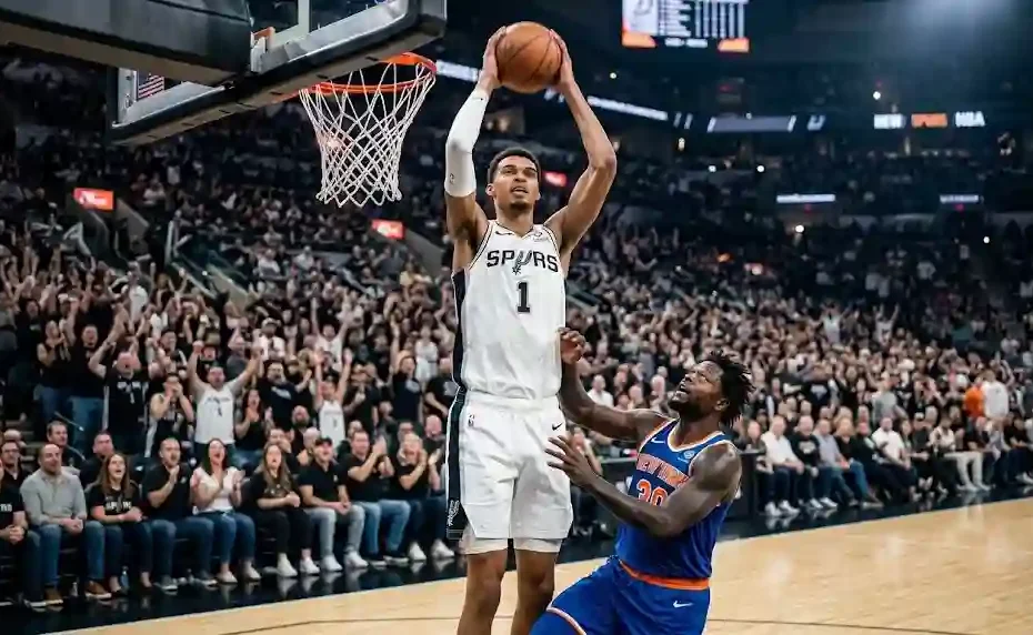 San Antonio Spurs center Victor Wembanyama going up for a two-handed dunk above a New York Knicks defender in front of a packed stadium crowd.