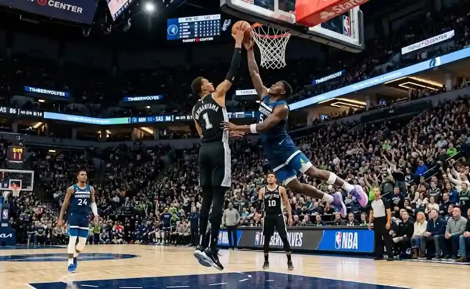 A Minnesota Timberwolves player jumping high to score at the rim against a San Antonio Spurs defender during an intense NBA game inside a crowded arena.