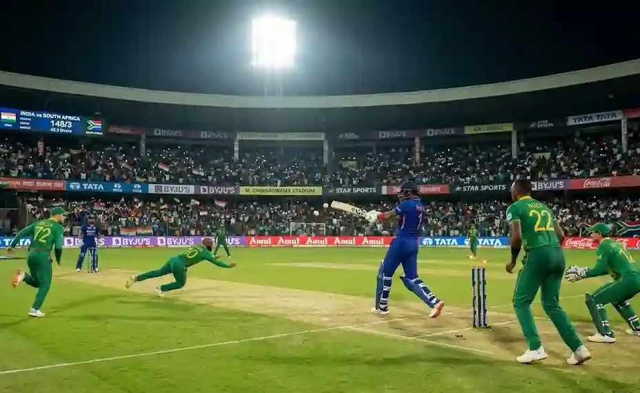 Photorealistic wide-angle shot of an India vs South Africa cricket match under stadium floodlights, featuring an Indian batsman hitting a drive against South African fielders.